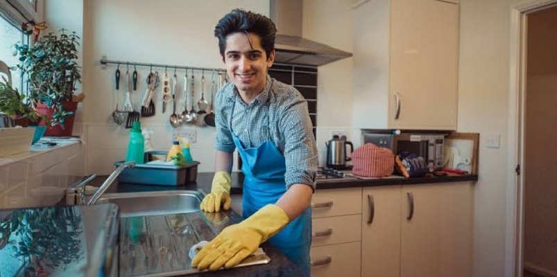 A young man wearing an apron and gloves, cleaning a kitchen surface and smiling.