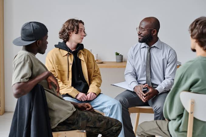 A mentor in conversation with a small group of young men seated in a bright room.